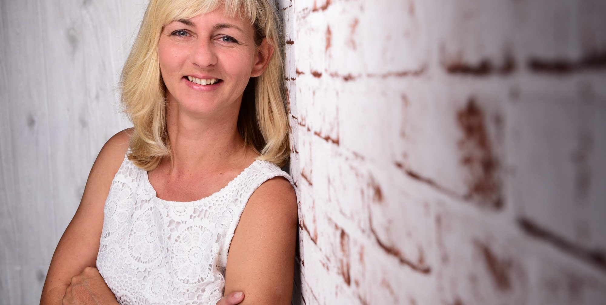 Porträt von Doreen Unglaube vor einer weiß-roten Backsteinwand Porträt einer lächelnden Frau mit blonden Haaren. Sie trägt ein weißes Oberteil und lehnt seitlich an einer hellen Backsteinwand.