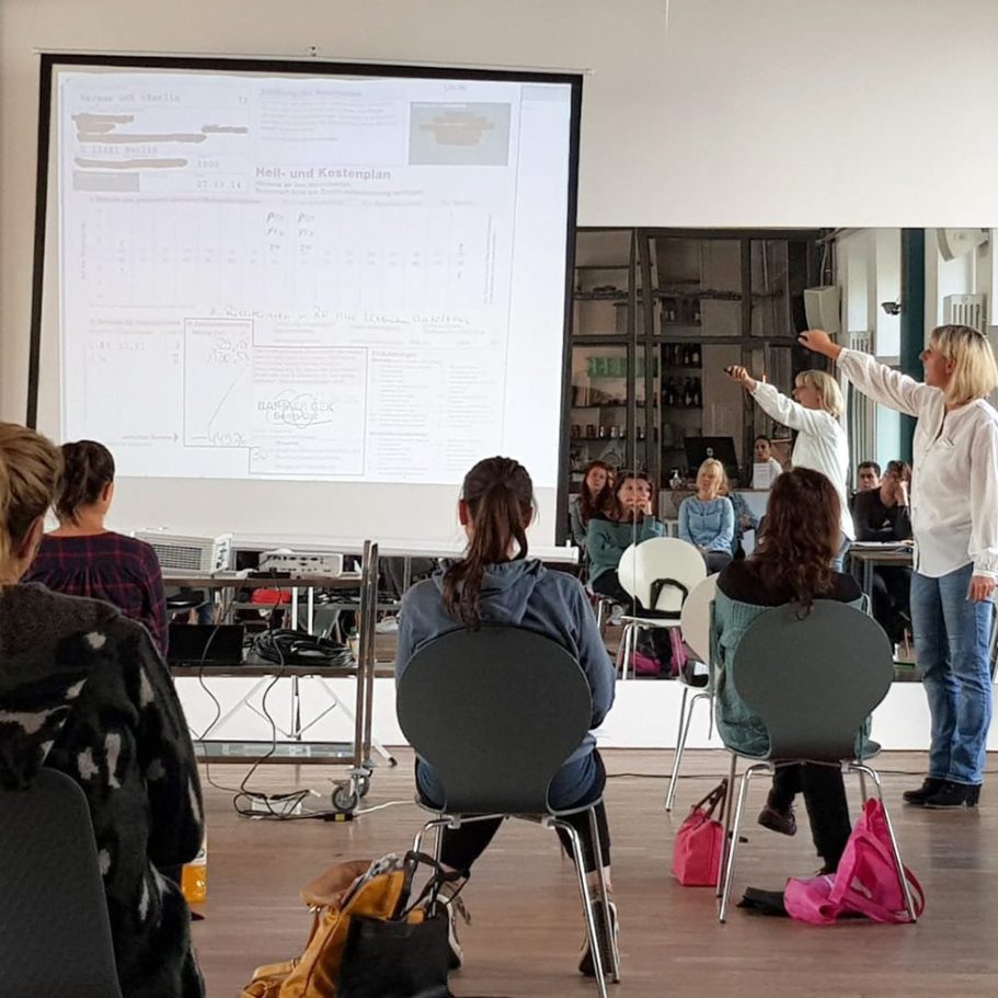 Seminar zur Abrechnung von Heil- und Kostenplänen für Zahnärzte Eine Referentin steht vor einer Leinwand und erklärt einer Gruppe von Teilnehmenden einen projizierten Heil- und Kostenplan.